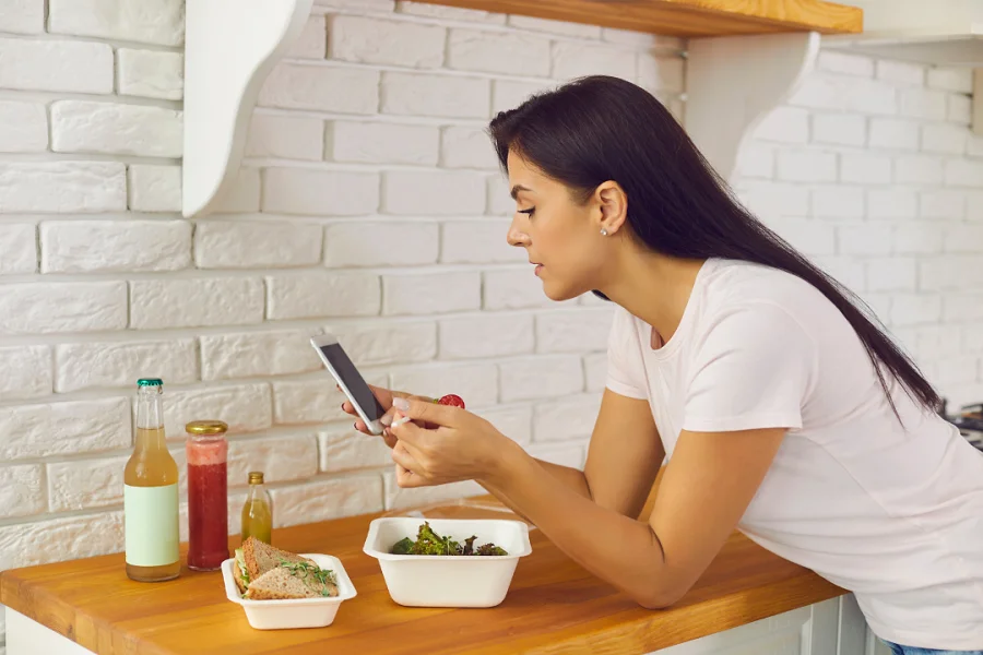 A woman looking at her phone while eating food and standing in the kitchen